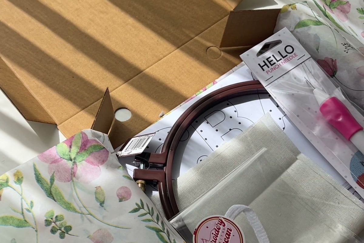 꽃무늬 포장지 위에 수틀, 원단, 알파벳 도안, 펀치 니들, 키링 고리가 담긴 개봉한 DIY 키트 상자